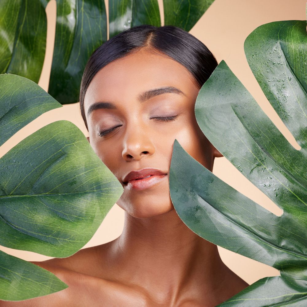 Una chica con una piel cuidada detrás de hojas de Monstera.