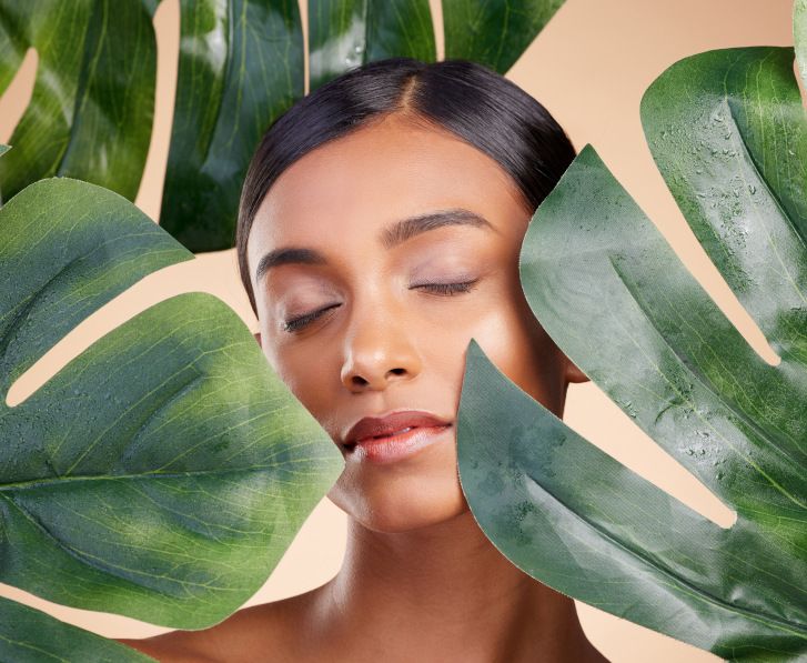 Una chica con una piel cuidada detrás de hojas de Monstera.